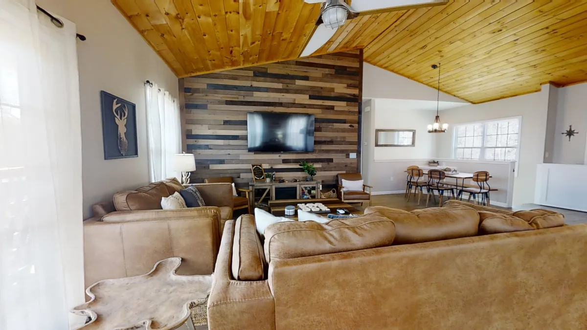 The image captures an open-concept living space with a large sectional sofa, wooden accent wall, flat-screen TV, and a dining area with chairs. Ample natural light floods through the windows, creating a welcoming and comfortable atmosphere ideal for both relaxation and entertaining.