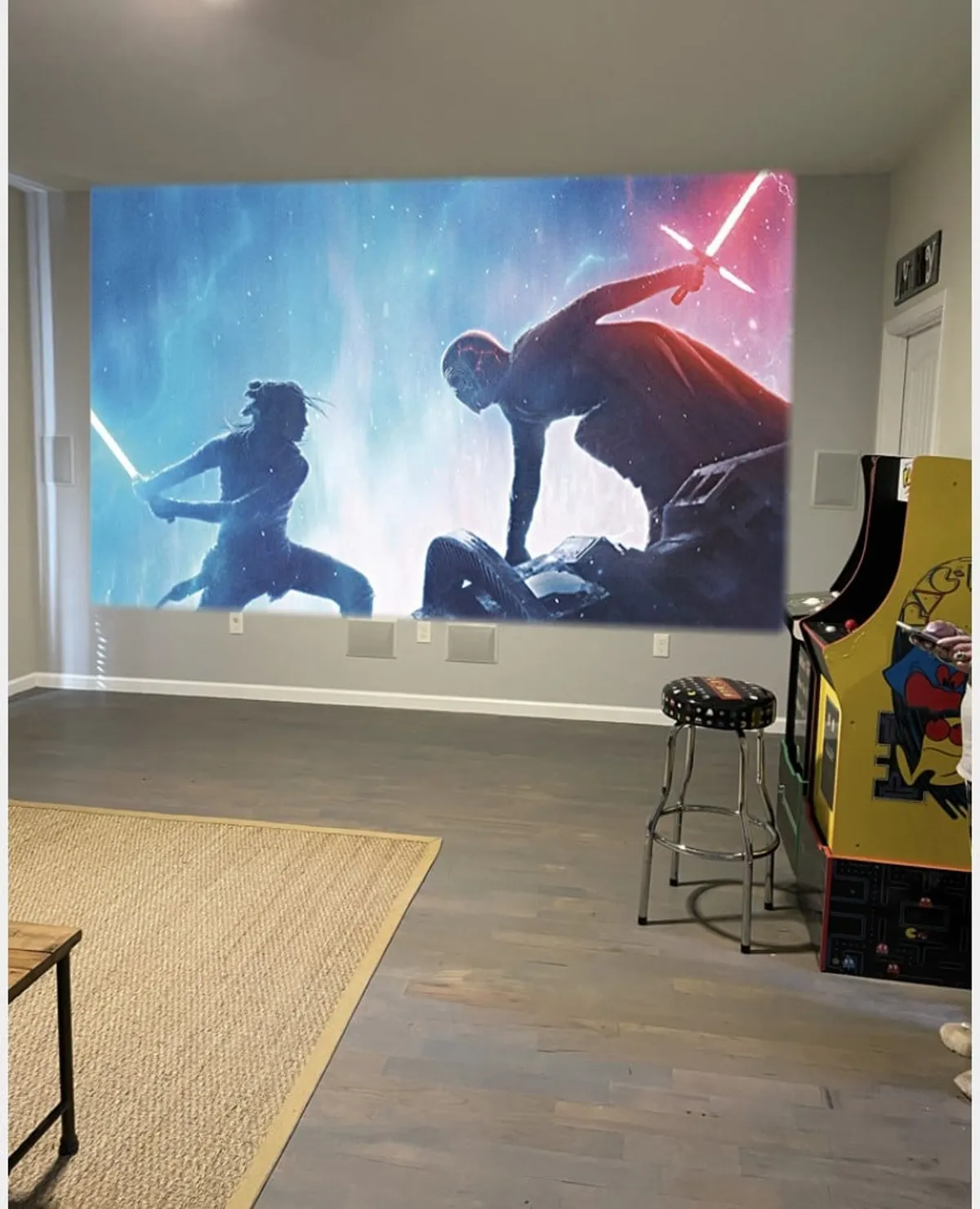 A vibrant interior game room featuring a large mural of a lightsaber duel, an arcade machine, and a bar stool. The space is energetic and playful, designed for gaming, movie nights, and socializing, making it ideal for families with kids and adults seeking fun indoor activities.
