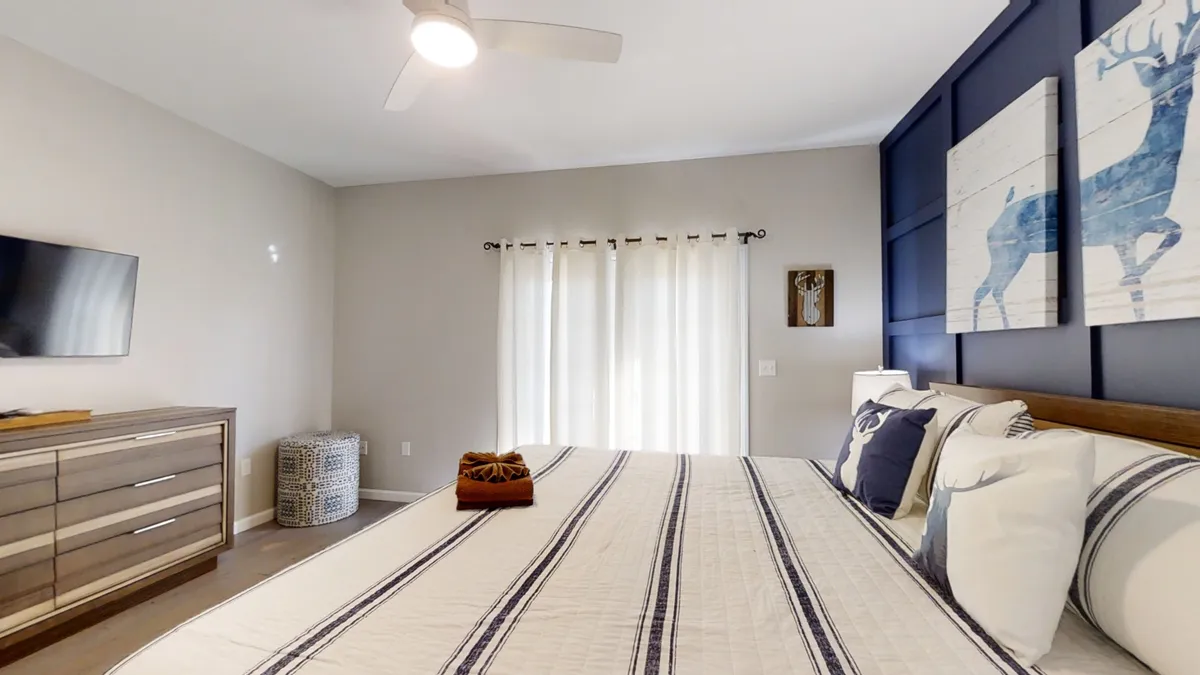 A serene bedroom with striped bedding set against a backdrop of a large window draped with sheer curtains. Decorative wall art featuring deer enhances the nature-inspired theme, while a modern dresser and television complete the cozy setup. Natural light fills the space, suggesting inviting mornings and a calming connection to the outdoors.