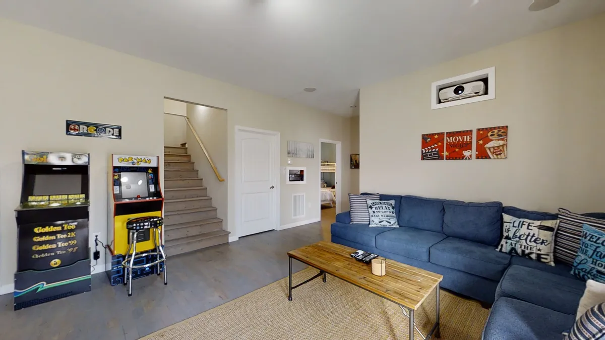 A spacious entertainment area with a plush blue sectional sofa, wooden coffee table, visible arcade machines, and a staircase leading to the upper level. The scene hints at a multifunctional space where people can relax, gather, and enjoy both games and movies.