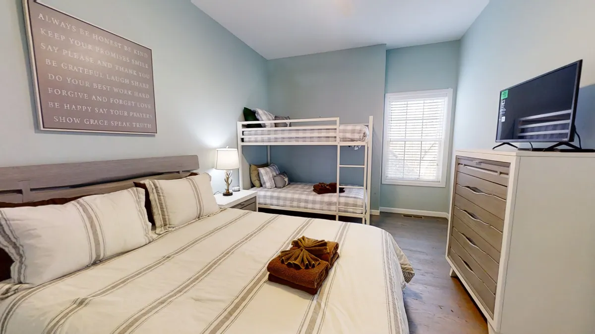 A spacious bedroom featuring a large bed adorned with decorative pillows and a cozy bunk bed. The room is thoughtfully arranged, with a dresser topped by a television and a framed inspirational quote on the wall, creating a family-friendly and inviting environment.