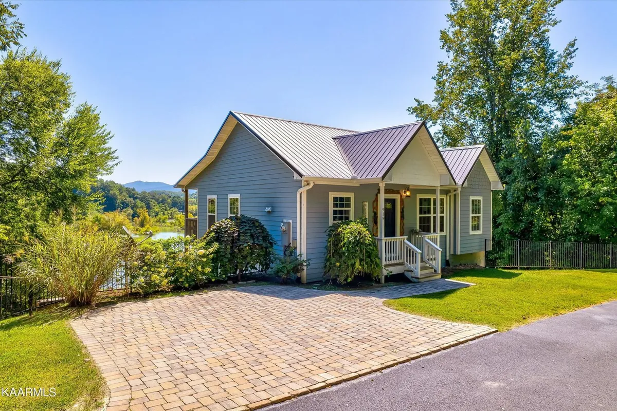 A welcoming front porch with a landscaped garden, paved driveway, and a glimpse of the metal roof. The porch overlooks a serene natural setting, hinting at the beauty of the surrounding area and the inviting atmosphere of the property. The outdoor furniture on the porch suggests a place to relax after a day of adventure.