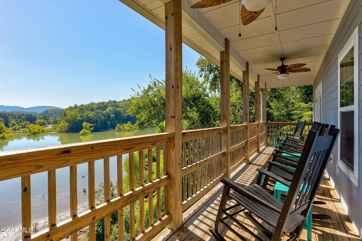 This exterior image features a deck outfitted with classic wooden rocking chairs, a sturdy wooden railing, and sweeping views of a serene river with mountains in the distance. The tranquil mood is enhanced by the suggestion of early morning mist or a dramatic sunset glow, making the outdoor space feel inviting and peaceful.