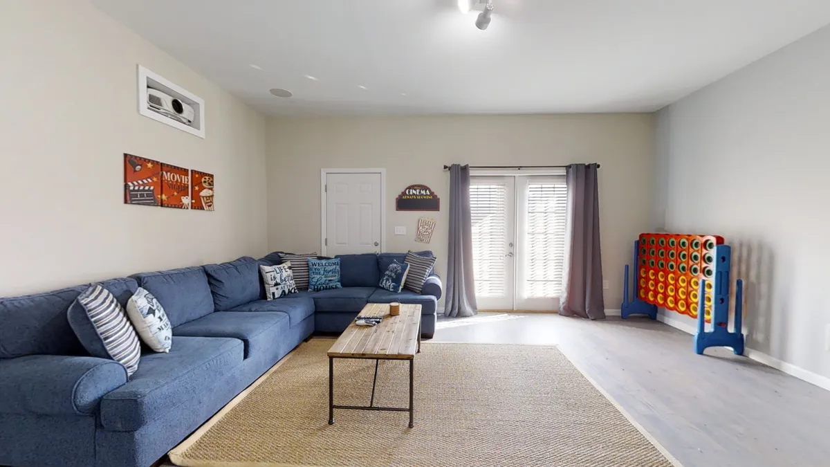 A spacious and casual living area with a blue sectional sofa, coffee table, and double doors leading outside. A colorful game board is set up, ready for group play. The room is bright, with a welcoming, playful ambiance suited for gatherings.